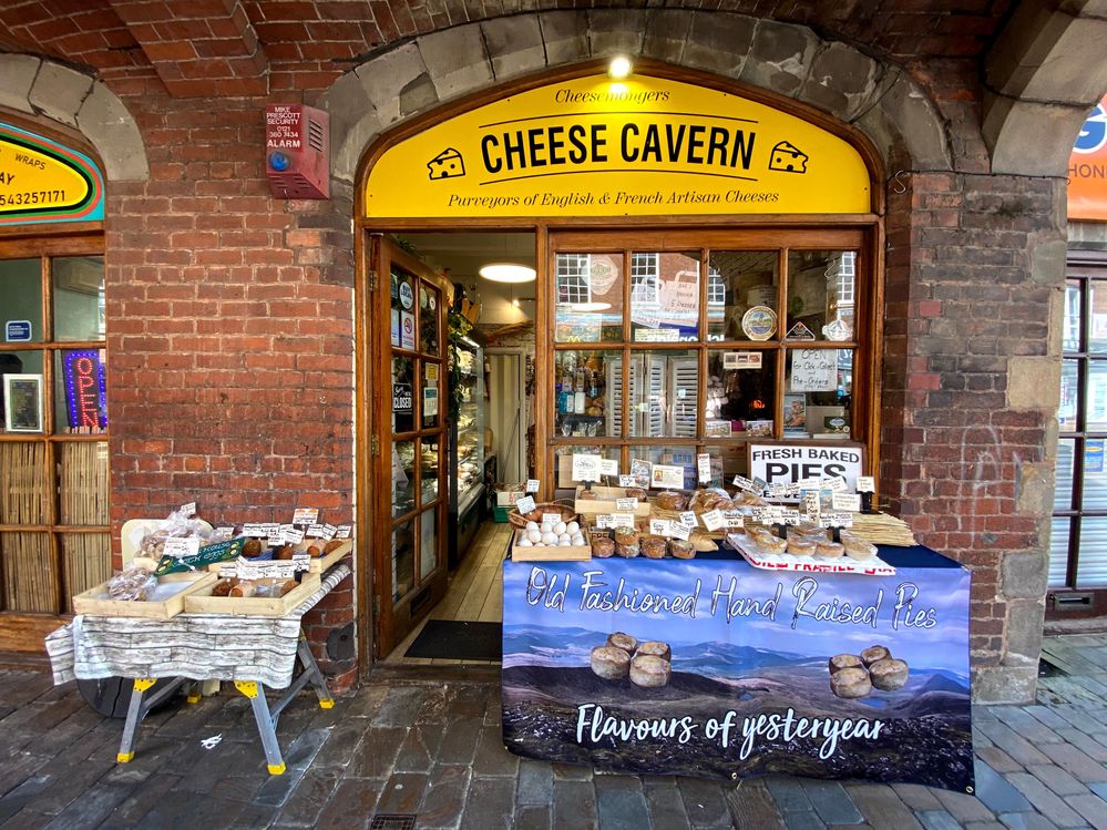 Caption: A storefront photo of the Cheese Cavern in Lichfield, UK, with the door open to show the inside and two tables with pastries on display in front. (Local Guide David C.)