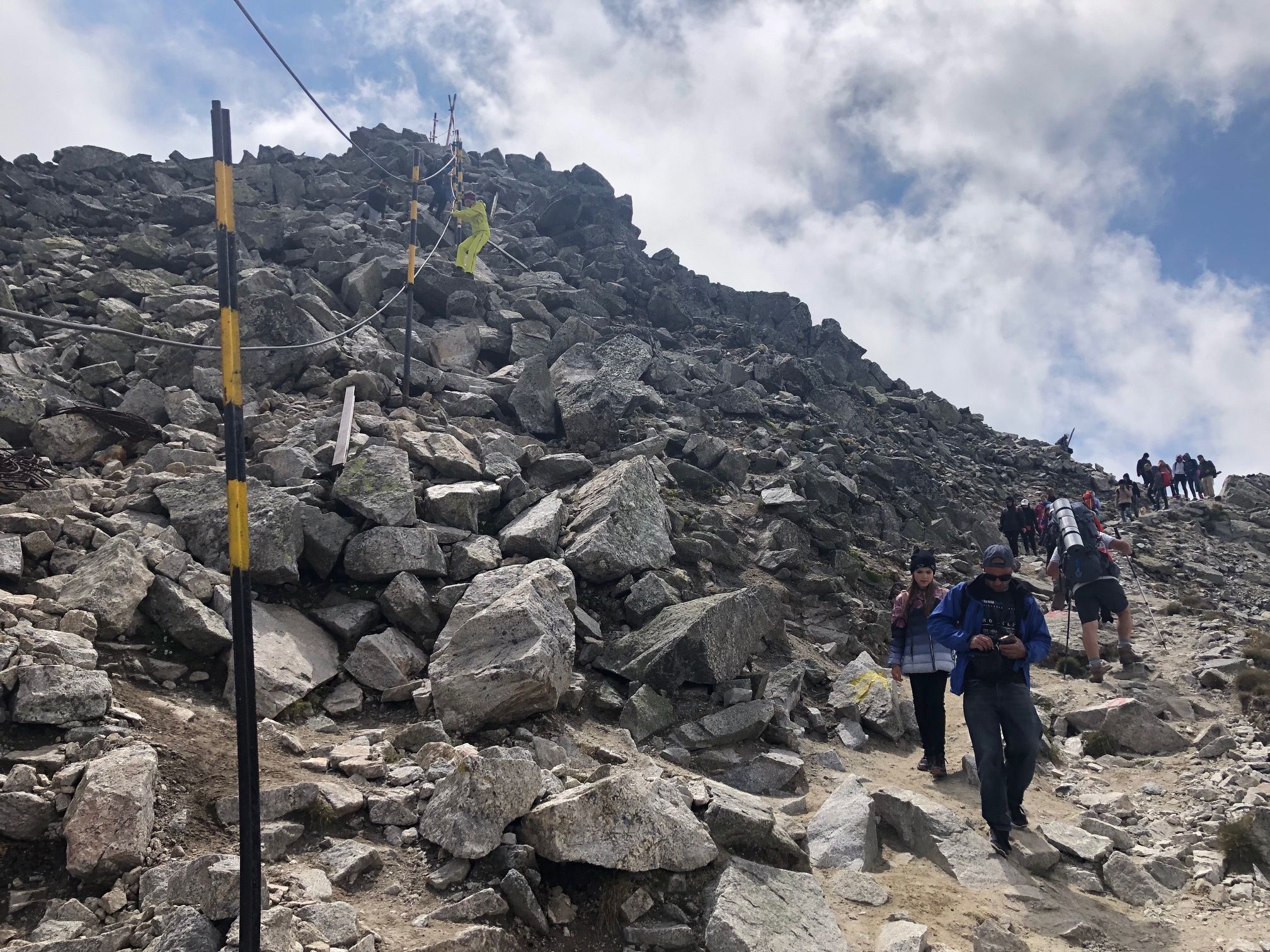 Caption: A photo showing the two paths that hikers can take on the last slope towards Musala Peak. On the left there are tall metal bars and metal cables fixed over large stones and on the right—a flatter path with sand and stones, with many people going up and down. (Local Guide @FelipePk)