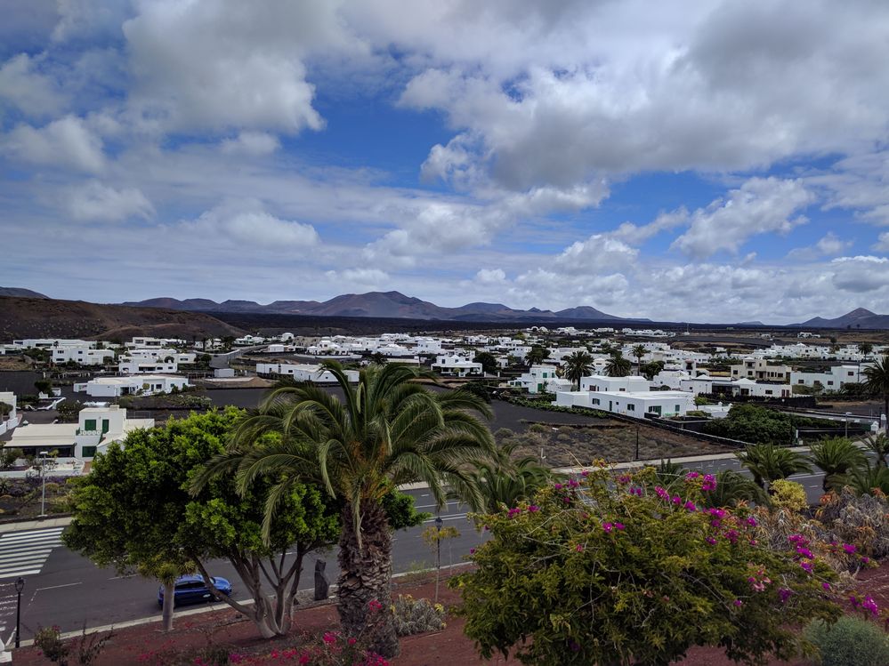 Caption: A photo of a city in Lanzarote, Canary Islands, with its white one-floor houses, green trees, flowers, and mountains in the background, taken on a partly cloudy day. (Local Guide @MoniDi)