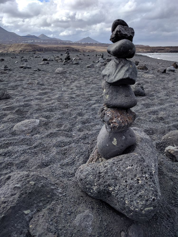 Caption: A photo of the black sand beach at Faro de Pechiguera, Lanzarote, with several rocks arranged in a small stone tower, the sea, and the mountains in the background. (Local Guide @MoniDi)