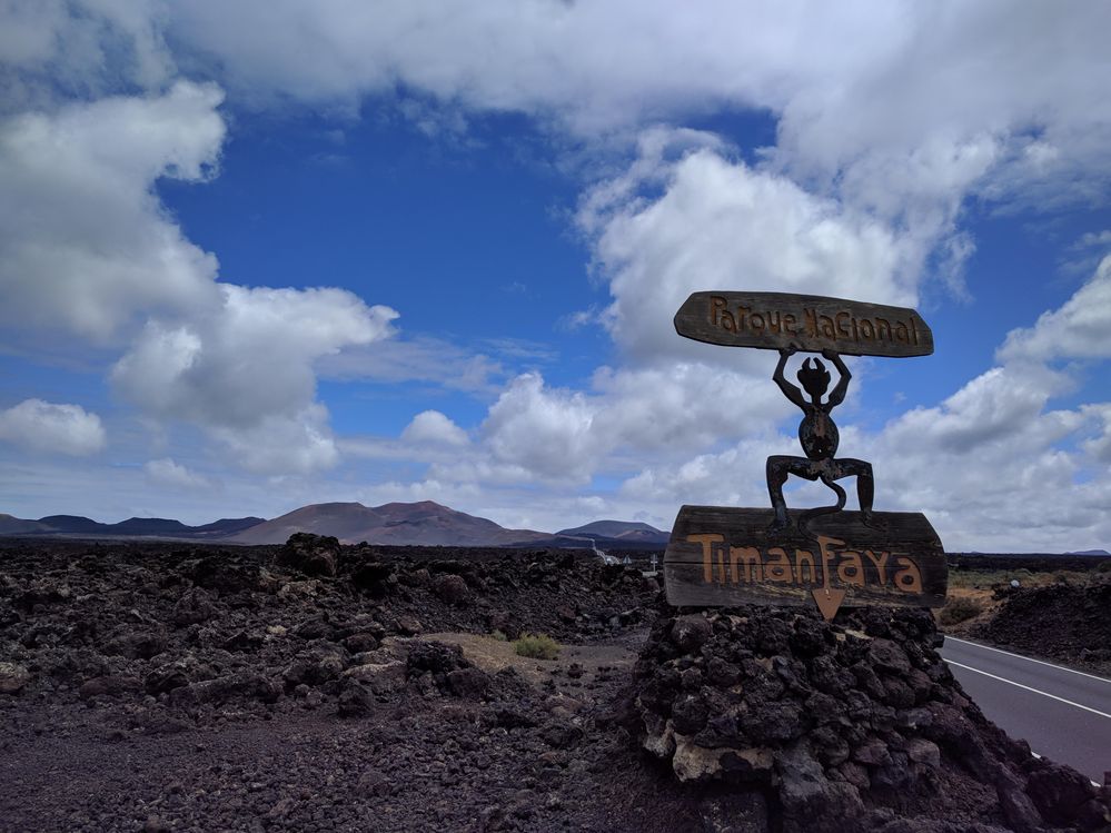 Caption: A photo of the wooden statue “El Diablo”, symbol of Timanfaya National Park, showing the figure of a devil, stepping on a wooden plate with the name “Timanfaya” and holding another name plate. The volcanic scenery of the park can be seen in the background. (Local Guide @MoniDi)