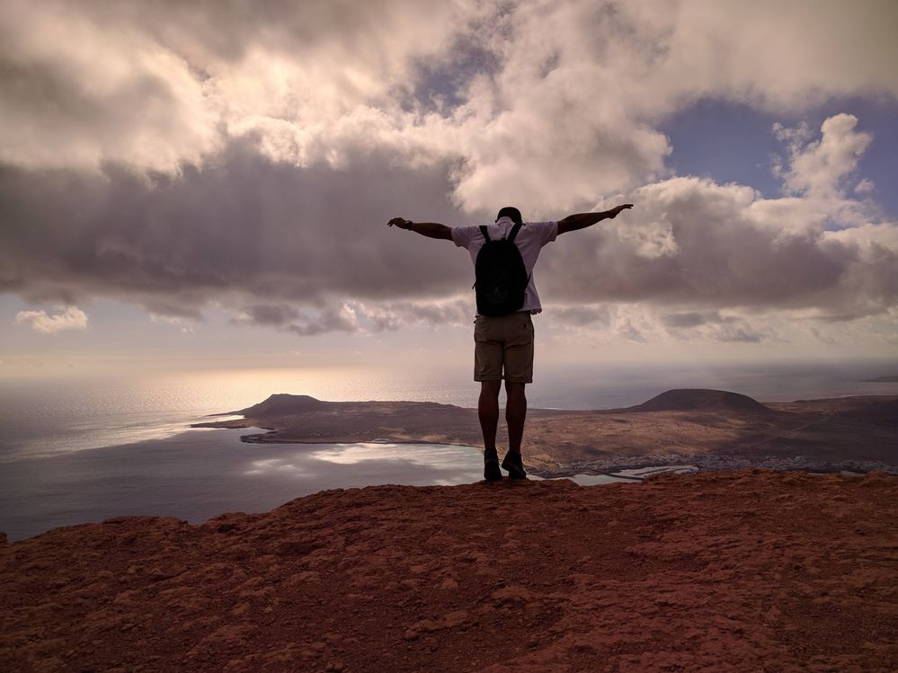 Caption: A photo of a man standing on the edge of a cliff at Mirador del Rio, Lanzarote, with the neighbouring island visible the background. (Local Guide @MoniDi)