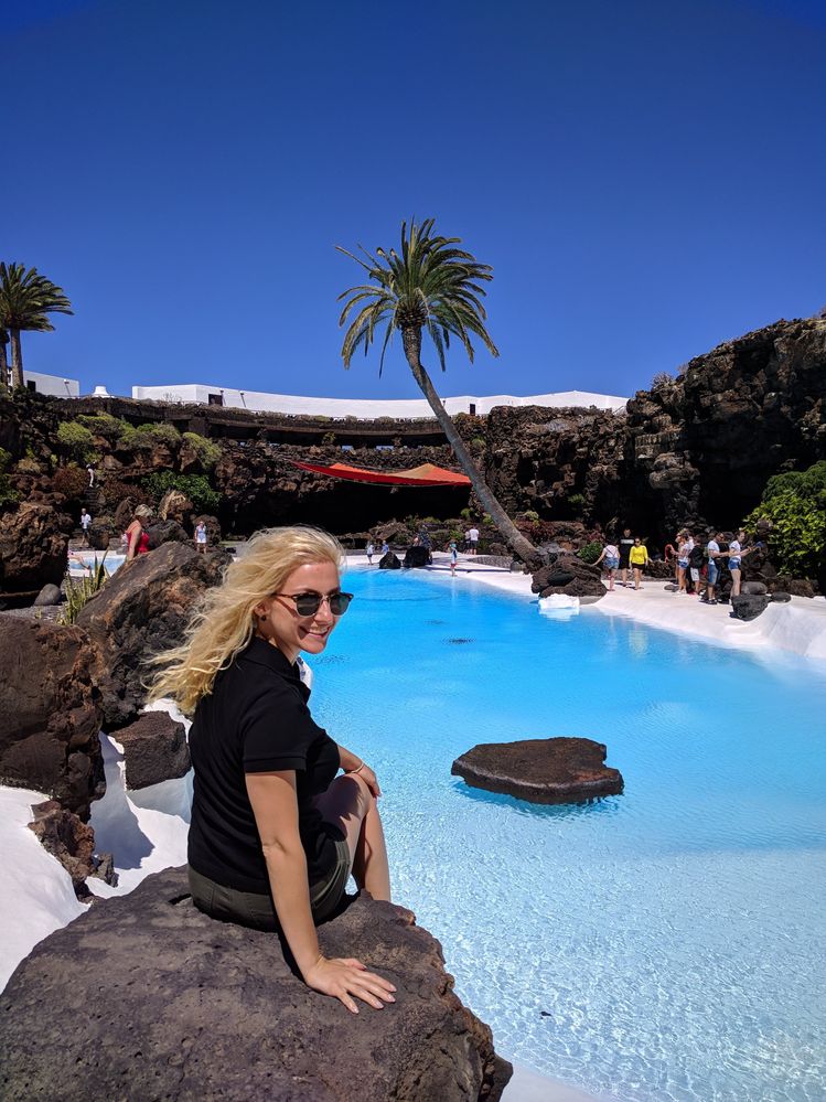 Caption: A photo of Google Moderator @MoniDi sitting on a rock in front of the bright blue pool at Jameos del Agua, Lanzarote. There are caves around the pool and a palm tree in the background. (Local Guide @MoniDi)