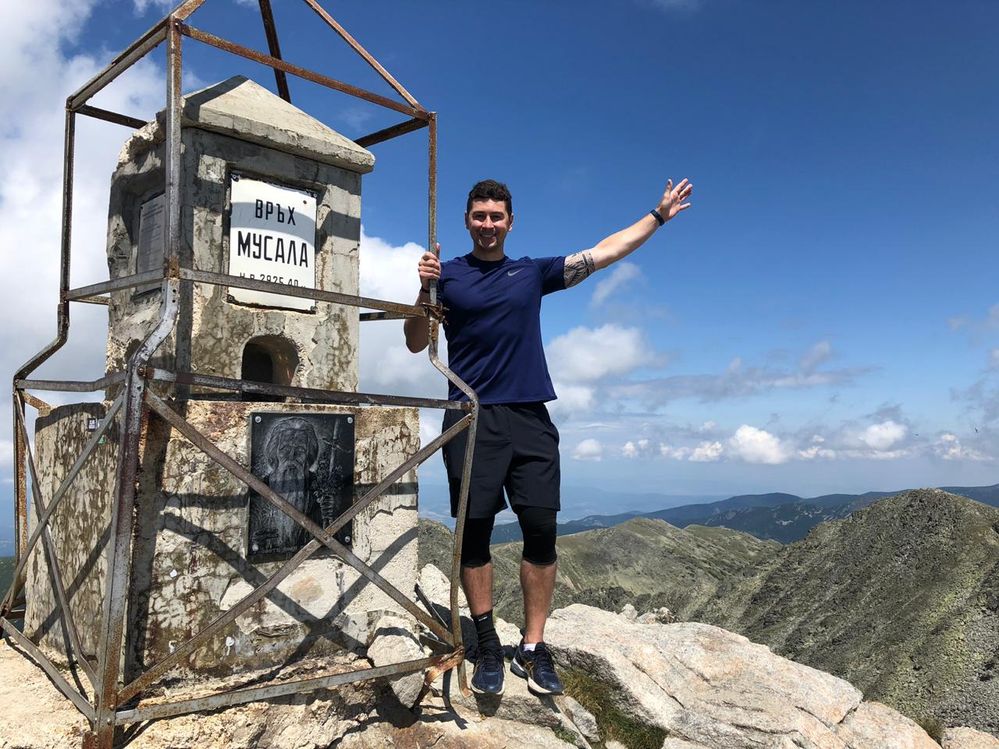 Caption: A photo of Google Moderator @FelipePk smiling and posing next to the stone monument on top of Musala Peak which has a metal plate with the name and the height of the peak and an image of a saint. (Local Guide @FelipePk)